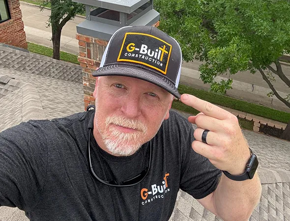 Man on a rooftop wearing a "G-Built Construction" cap and shirt, pointing to his hat. Background shows a chimney, trees, and street. Confident look.