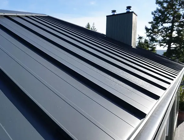 Sleek black Aluminum roof against a clear blue sky, featuring modern, clean lines. A tree and chimney add a touch of nature and structure.