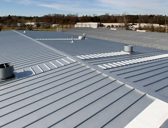 Aerial view of a large, metallic industrial roof with gray panels and small vents, set against a backdrop of distant trees and buildings under a blue sky.