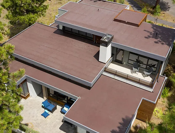 Aerial view of a modern flat-roofed house with brown roofing, surrounded by trees. Features a patio with blue outdoor furniture, exuding a serene, contemporary vibe.