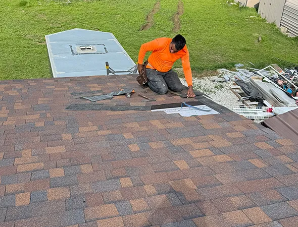 A person in an orange shirt repairs shingles on a roof, surrounded by tools and materials. The background shows a grassy yard and scattered items.