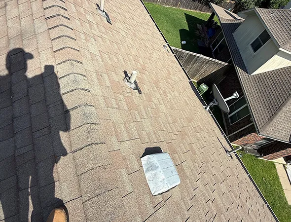 Shadow of a person in a hat, standing on a shingled roof. Nearby are rooftop fixtures, with a view of a house and green yard below. Bright, sunny day.
