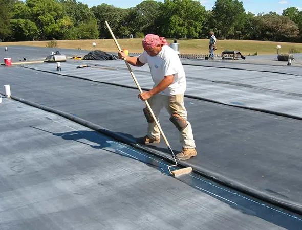 A worker wearing a red bandana and white shirt applies a coating on a flat roof with a roller. The surrounding area is tree-lined and sunny.