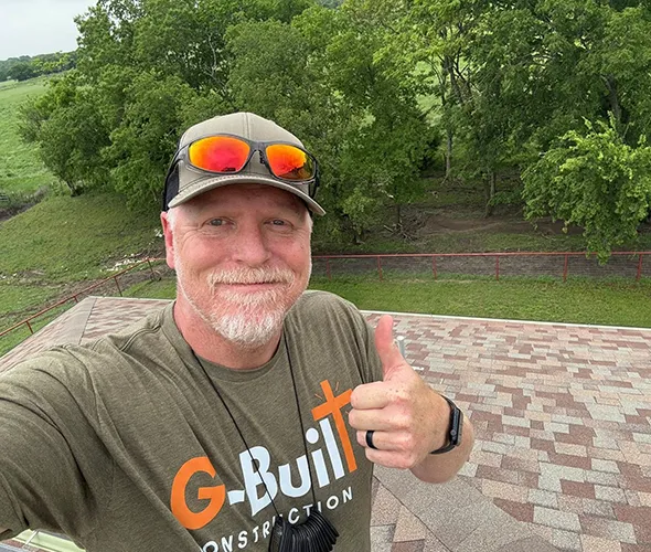 Man in a green "G-Built Construction" shirt gives a thumbs-up on a rooftop, smiling. He wears a cap and sunglasses, with trees in the background.